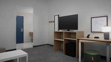 A hotel room with a flat-screen TV, a desk, a chair, a small table, a trash bin, and a green ottoman.
