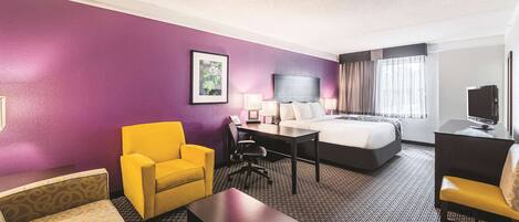 A hotel room with a purple wall, a bed, a desk with a TV, a yellow armchair, and a patterned sofa.