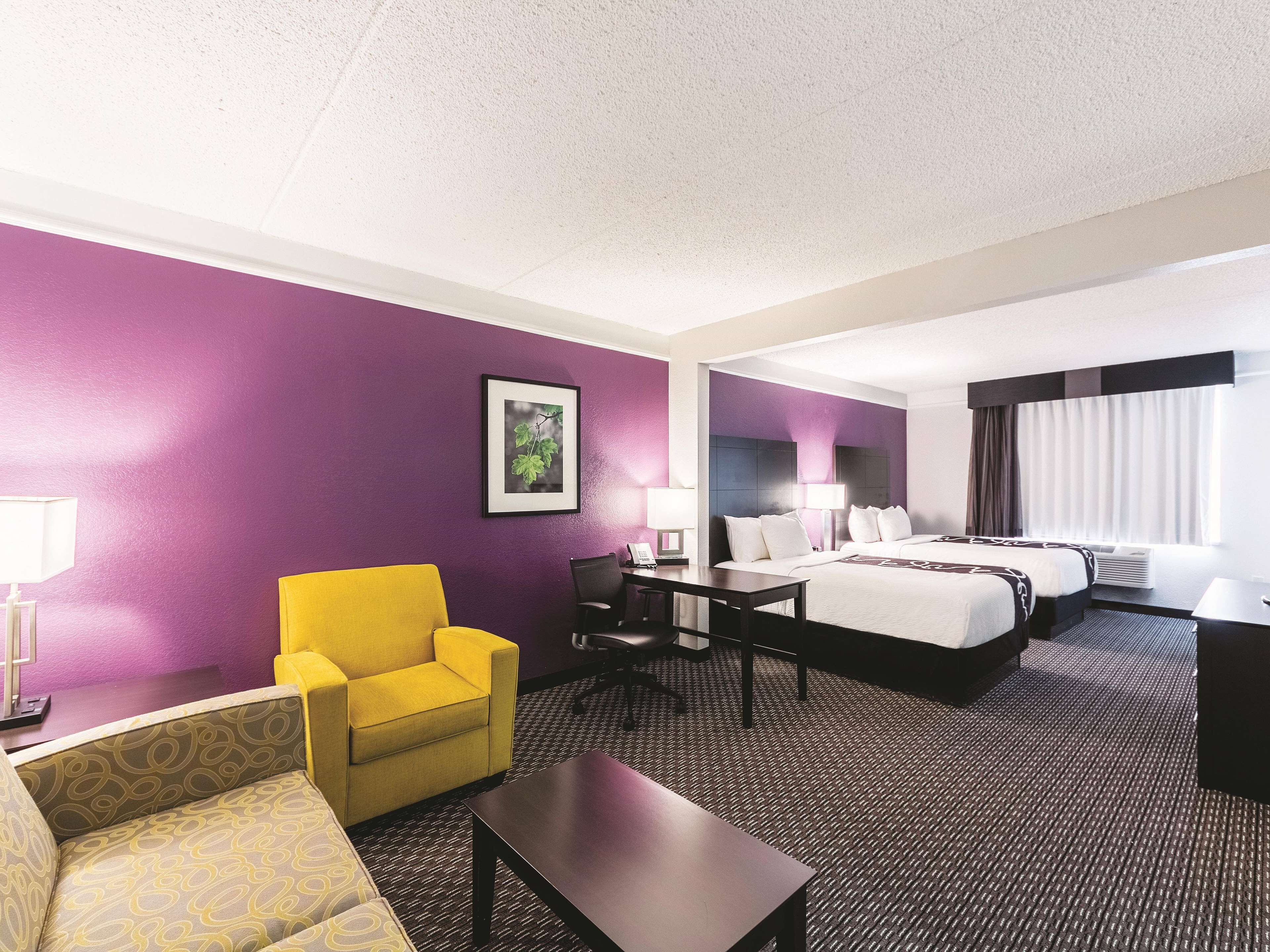 Ein Hotelzimmer mit violetter Wand, einem Bett, einem Schreibtisch, einem Stuhl und einem Sofa.