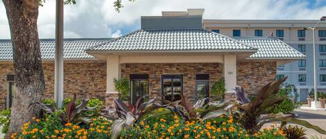 A building with a brick facade, a tiled roof, and a landscaped entrance with orange flowers and large plants.