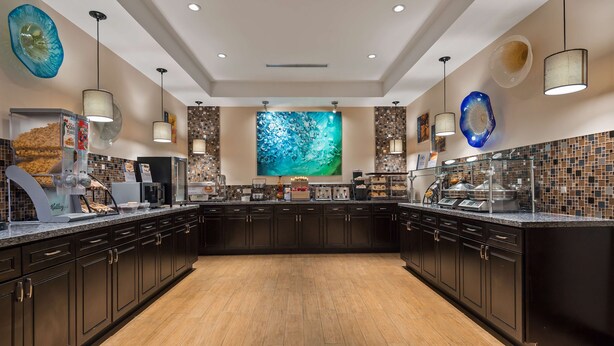 A modern kitchen with dark cabinetry, a wooden floor, and a large decorative wall mural. - Best Western Plus Hollywood/Aventura (Hallandale Beach)