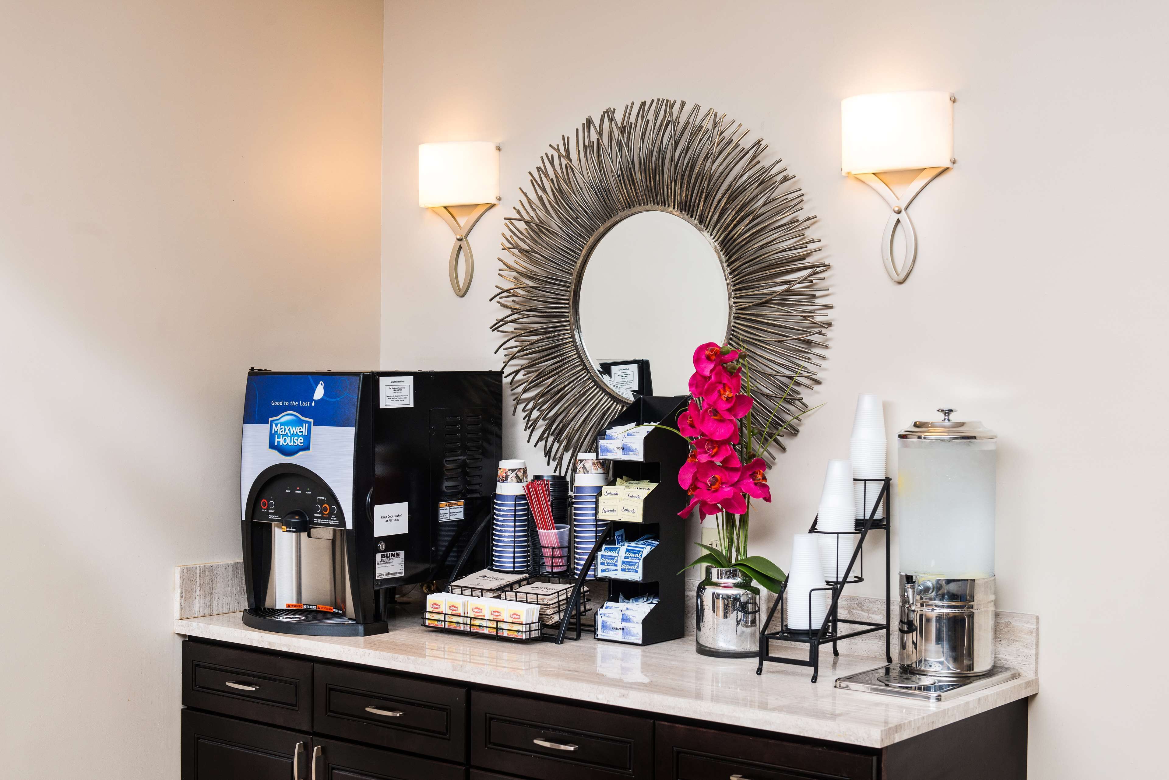 A hotel reception area with a coffee machine, a mirror, wall sconces, and a vase with flowers.