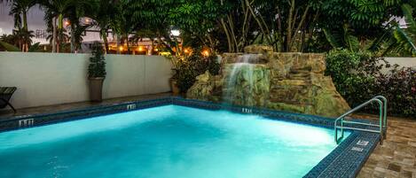 A rectangular pool with a waterfall feature, surrounded by lush greenery and a white wall.