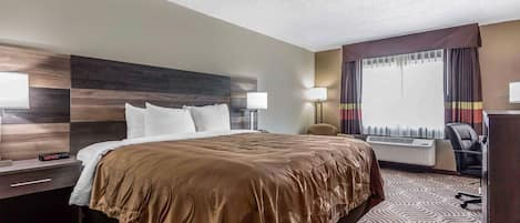 A hotel room with a large bed, a desk with a lamp, a chair, and a window with curtains.