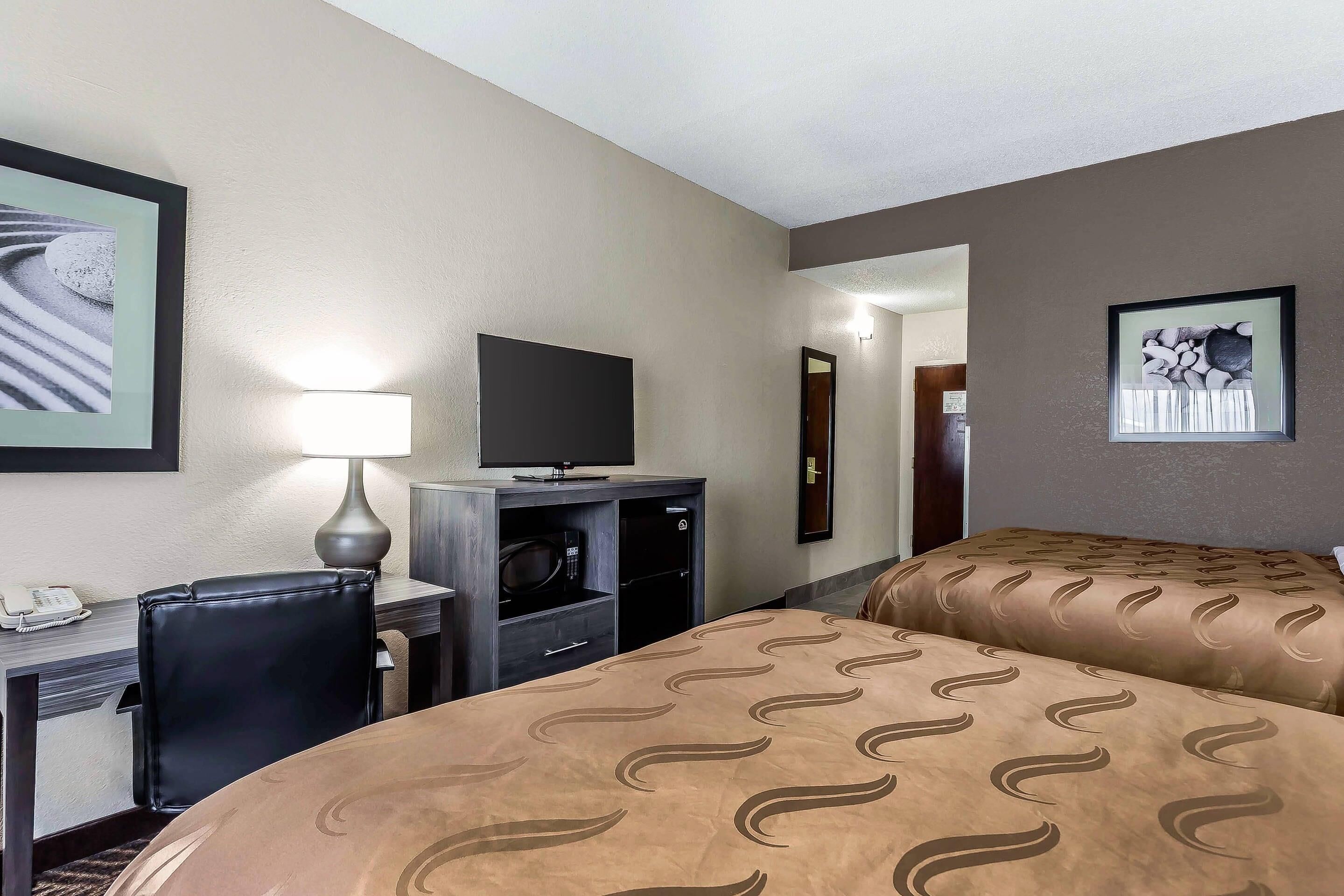 A hotel room with a desk, a television, a bed with a patterned bedspread, and a framed picture on the wall.