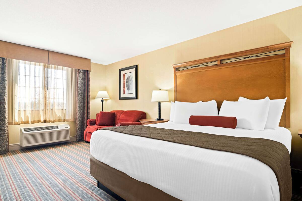 A hotel room with a large bed, a red chair, a desk with a lamp, and a window with curtains.