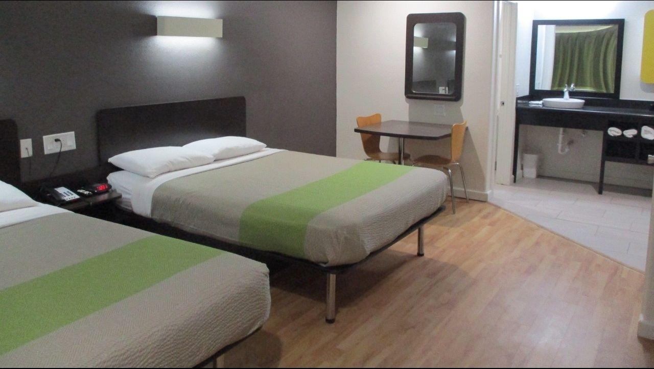 Quarto de hotel com duas camas de solteiro, uma mesa, uma cadeira, um espelho e banheiro com pia e espelho.