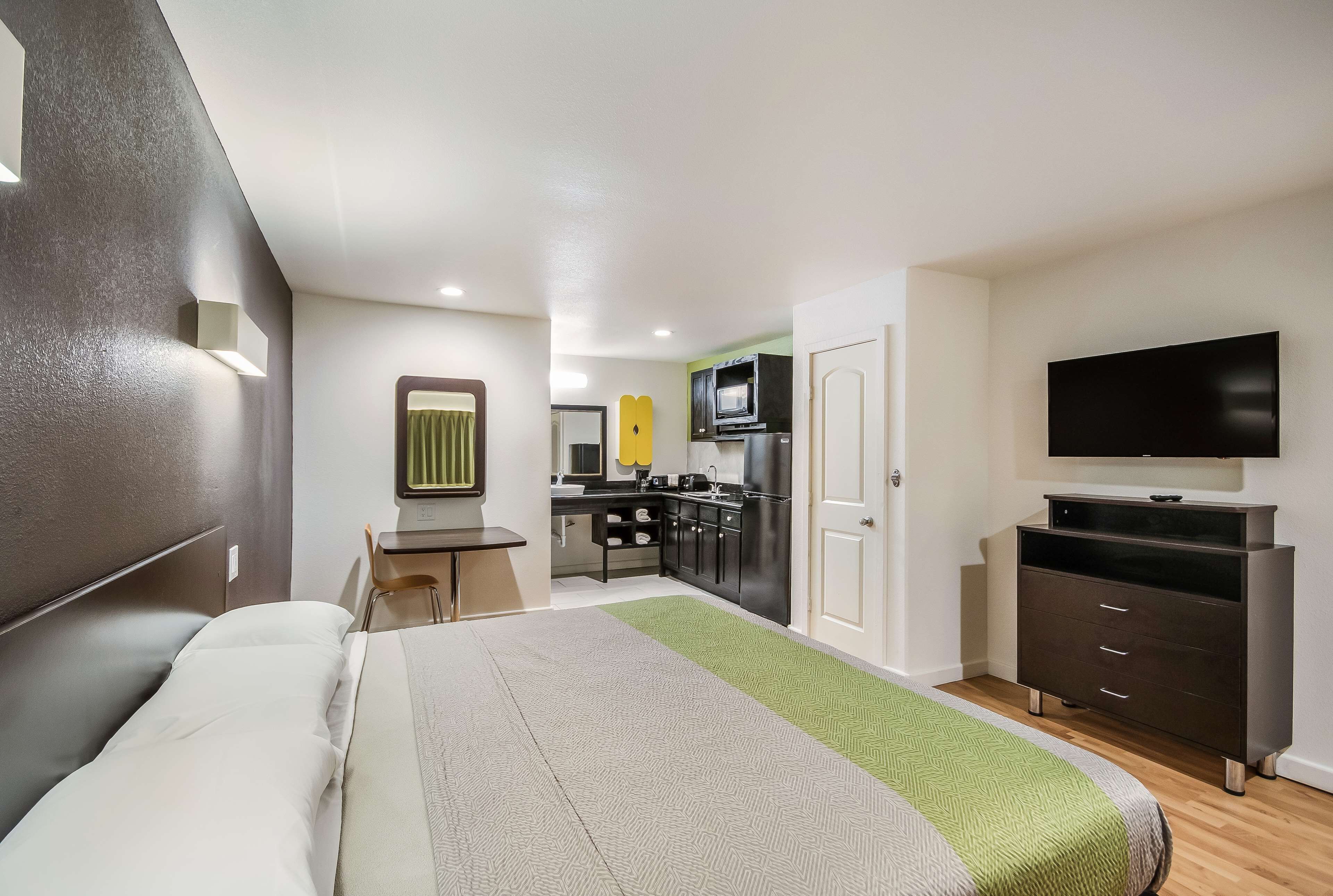 A hotel room with a bed, a desk, a kitchenette, and a TV.
