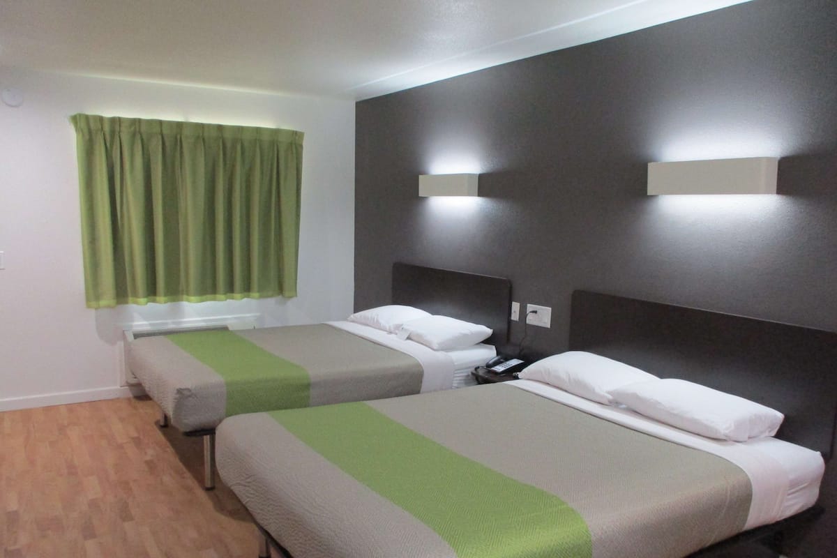 A hotel room with two beds, each with a green and grey bedspread, a bedside table with a lamp, and a window with green curtains.