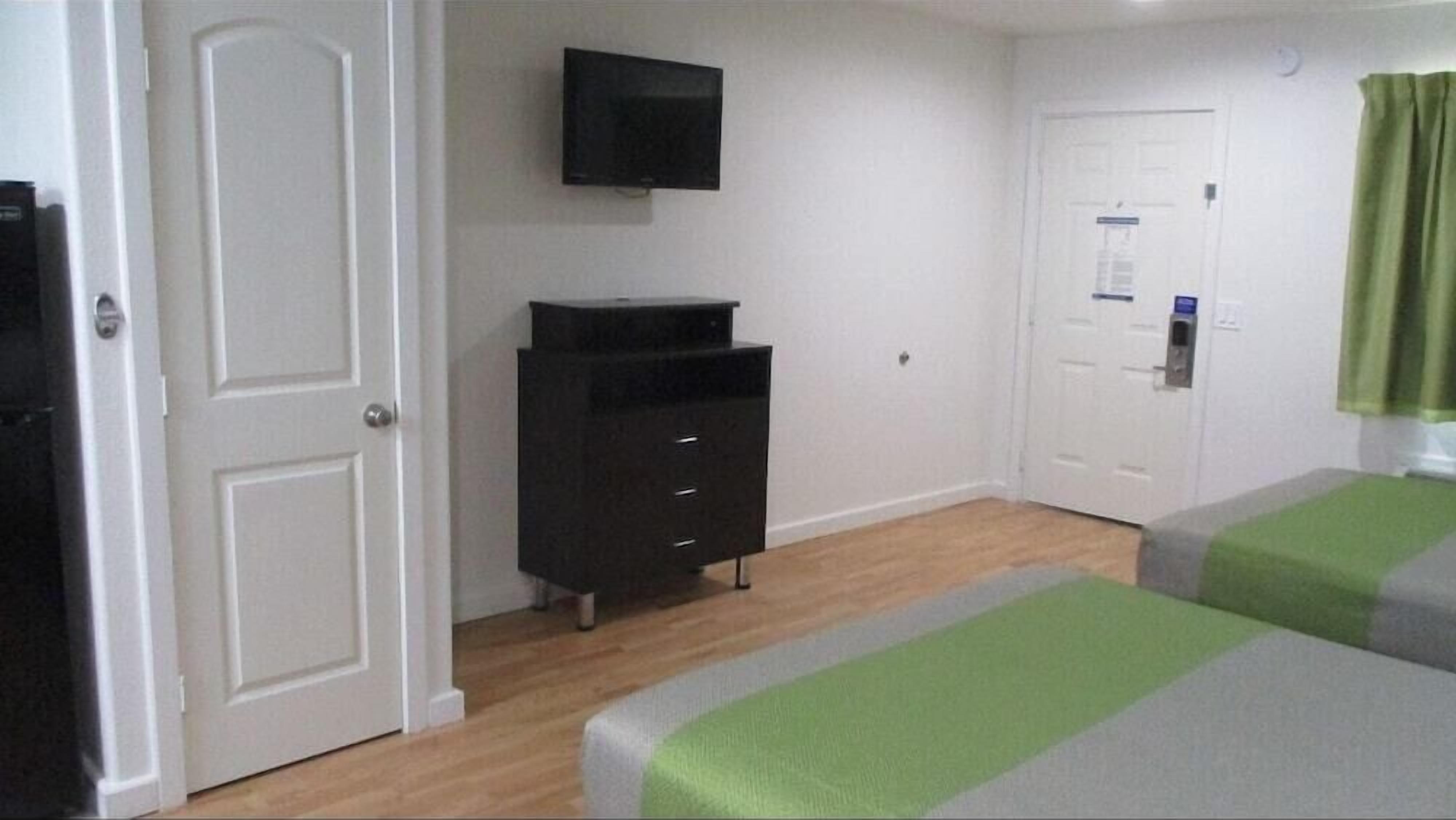 A room with a white door, a black cabinet, a television mounted on the wall, and two beds with green and gray bedding.