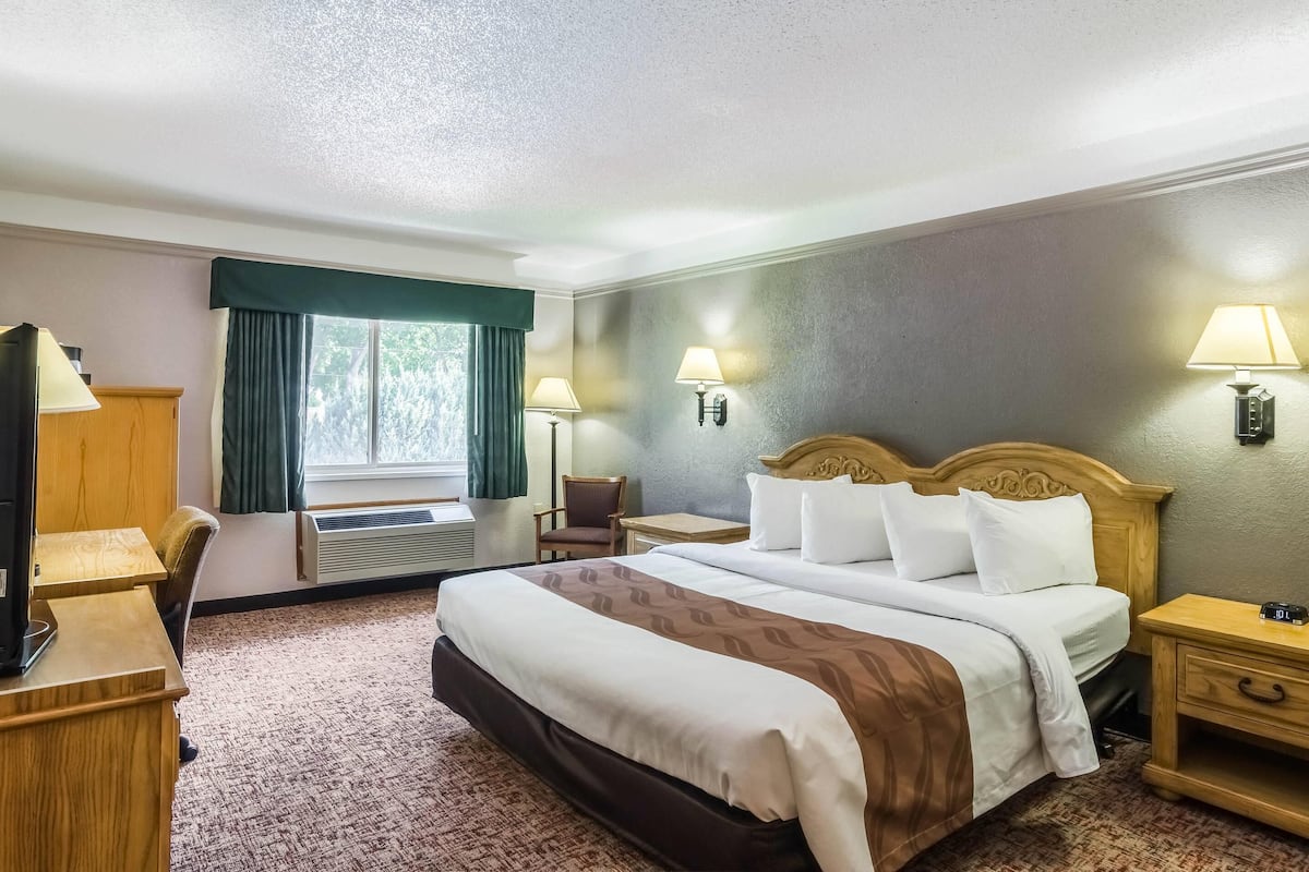A hotel room with a bed, bedside lamps, a desk, a chair, a window with curtains, and a thermostat.