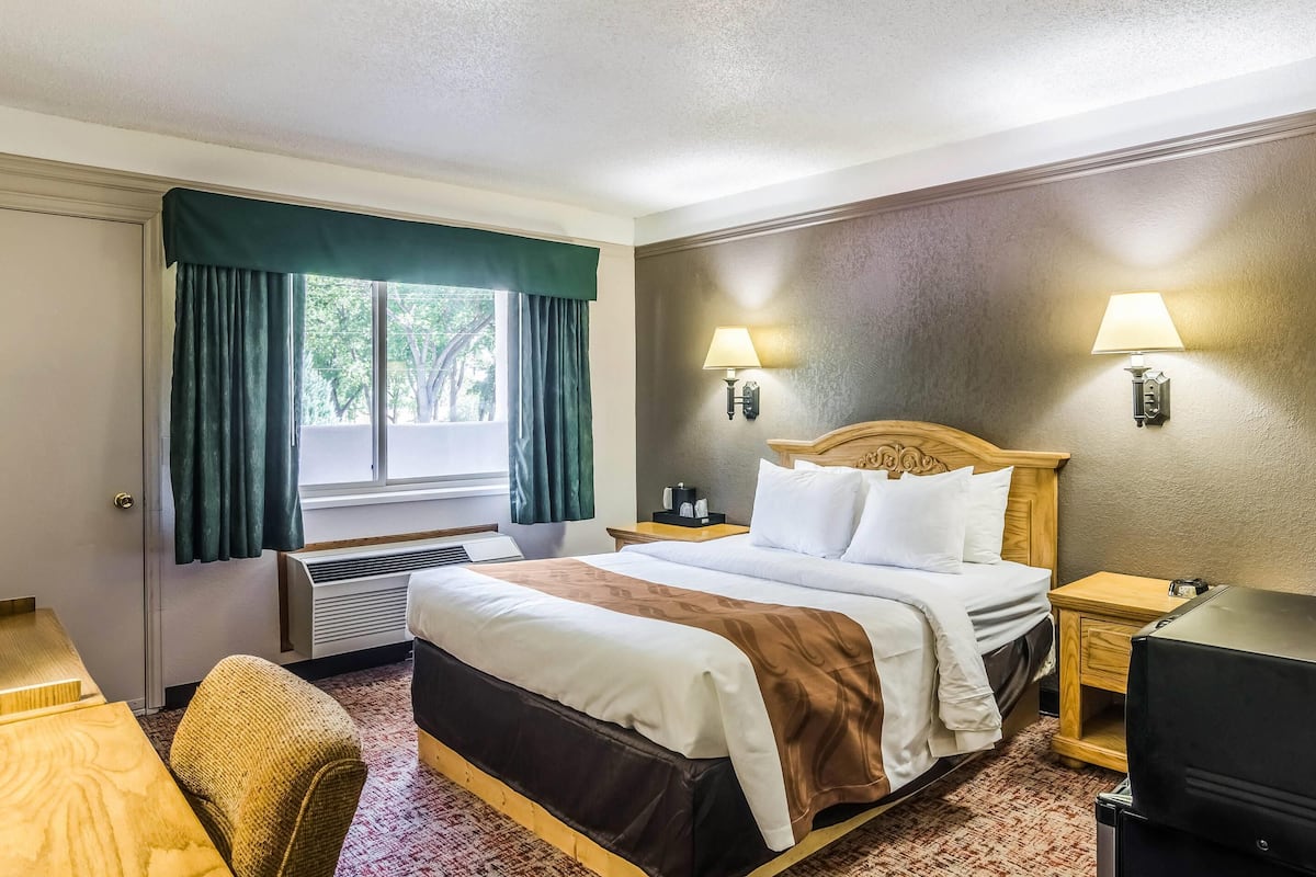 A hotel room with a bed, two bedside lamps, a wooden headboard, a desk, and a chair.