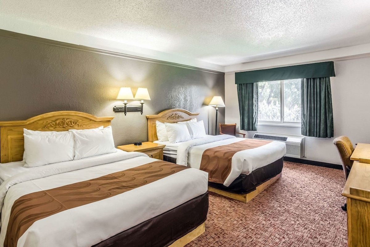 A hotel room with two beds, a desk, a chair, a window with curtains, and a wall-mounted lamp.