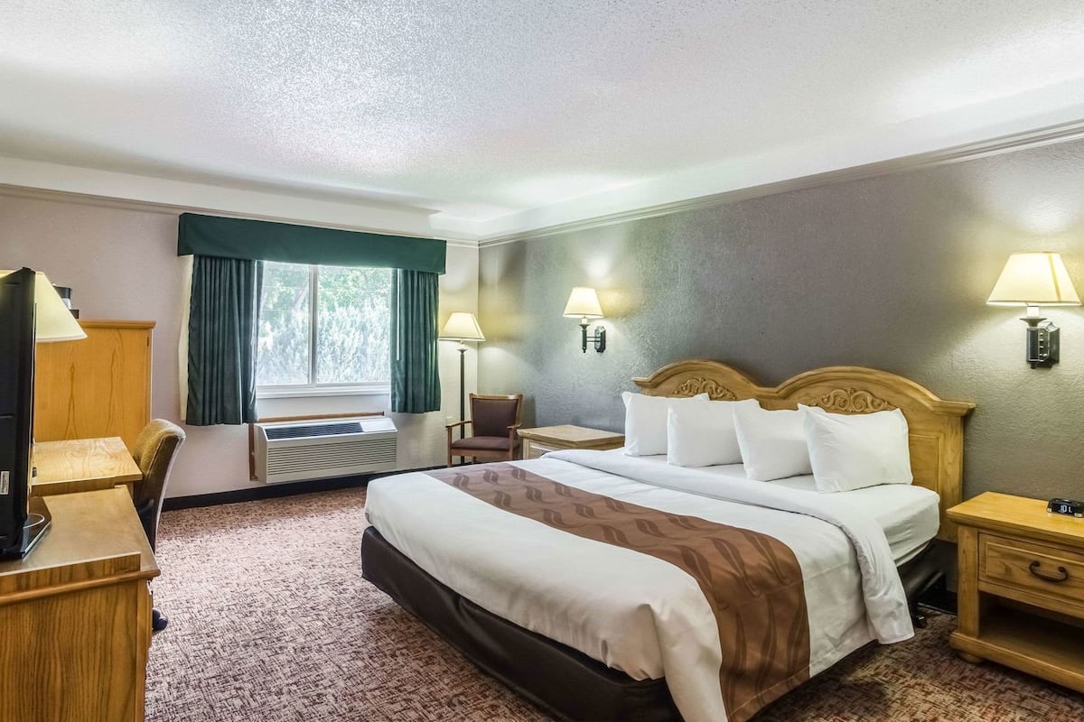 A hotel room with a bed, a desk, a chair, a TV, a lamp, and a clock.