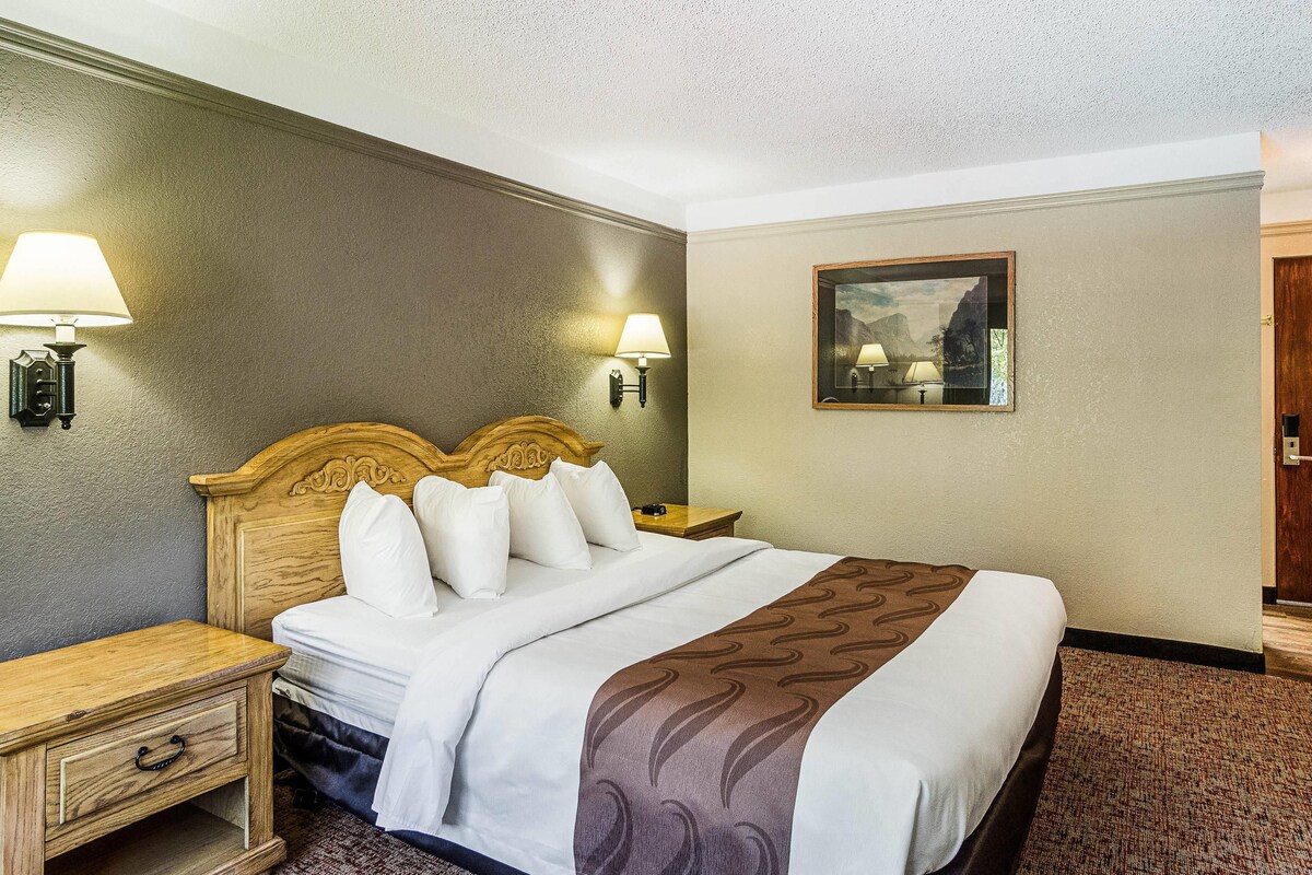 A hotel room with a wooden headboard, a bed with white bedding, two bedside lamps, a nightstand with a drawer, and a painting on the wall.
