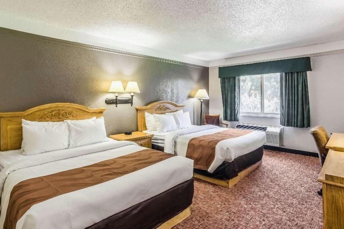 A hotel room with two beds, a desk, a chair, a window with curtains, and wall-mounted lamps.