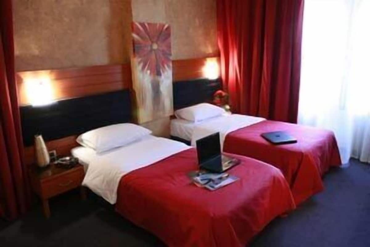 Habitación de hotel con dos camas, cada una con una colcha roja y almohadas blancas. Hay un portátil sobre la cama en primer plano.