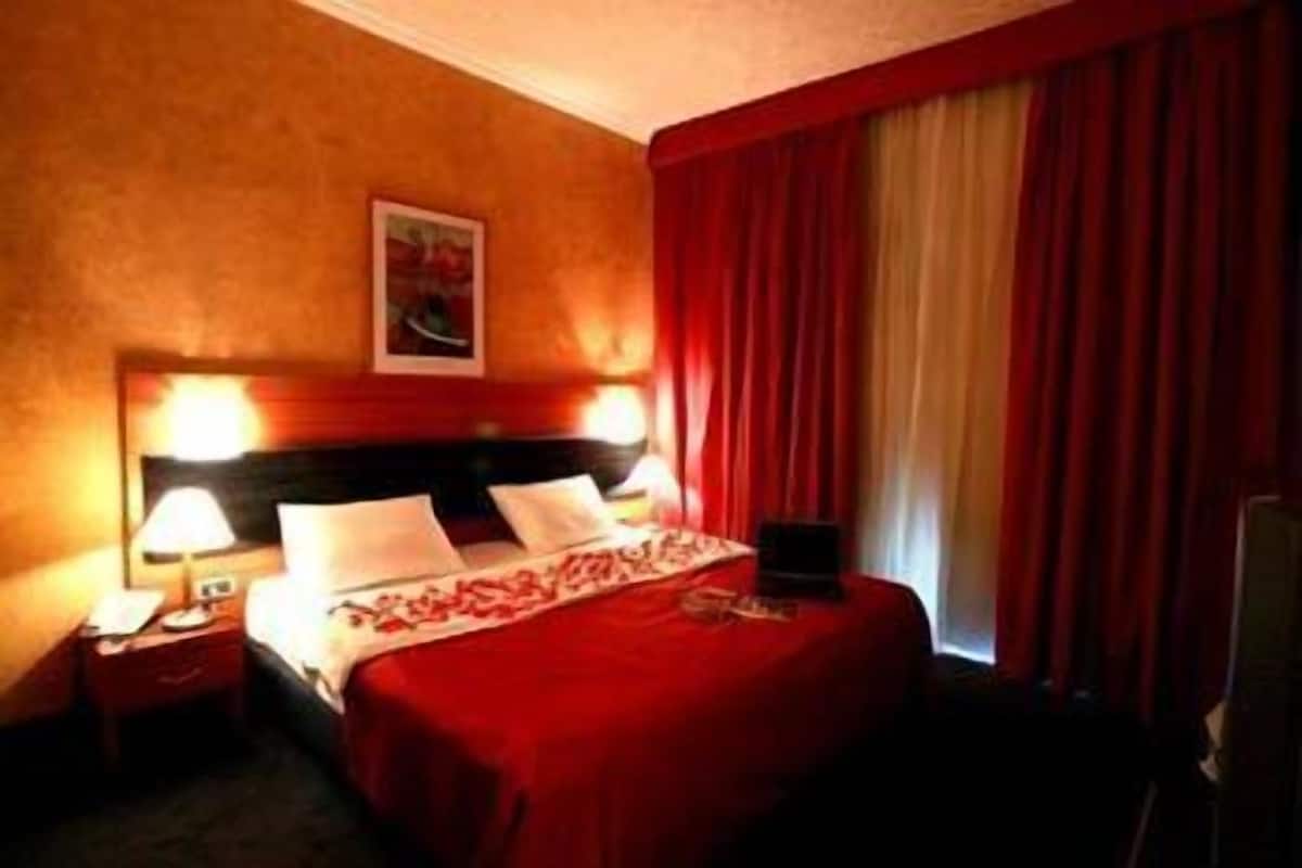 Una habitación de hotel con una cama grande, una mesita de noche con una lámpara y una ventana con cortinas rojas.