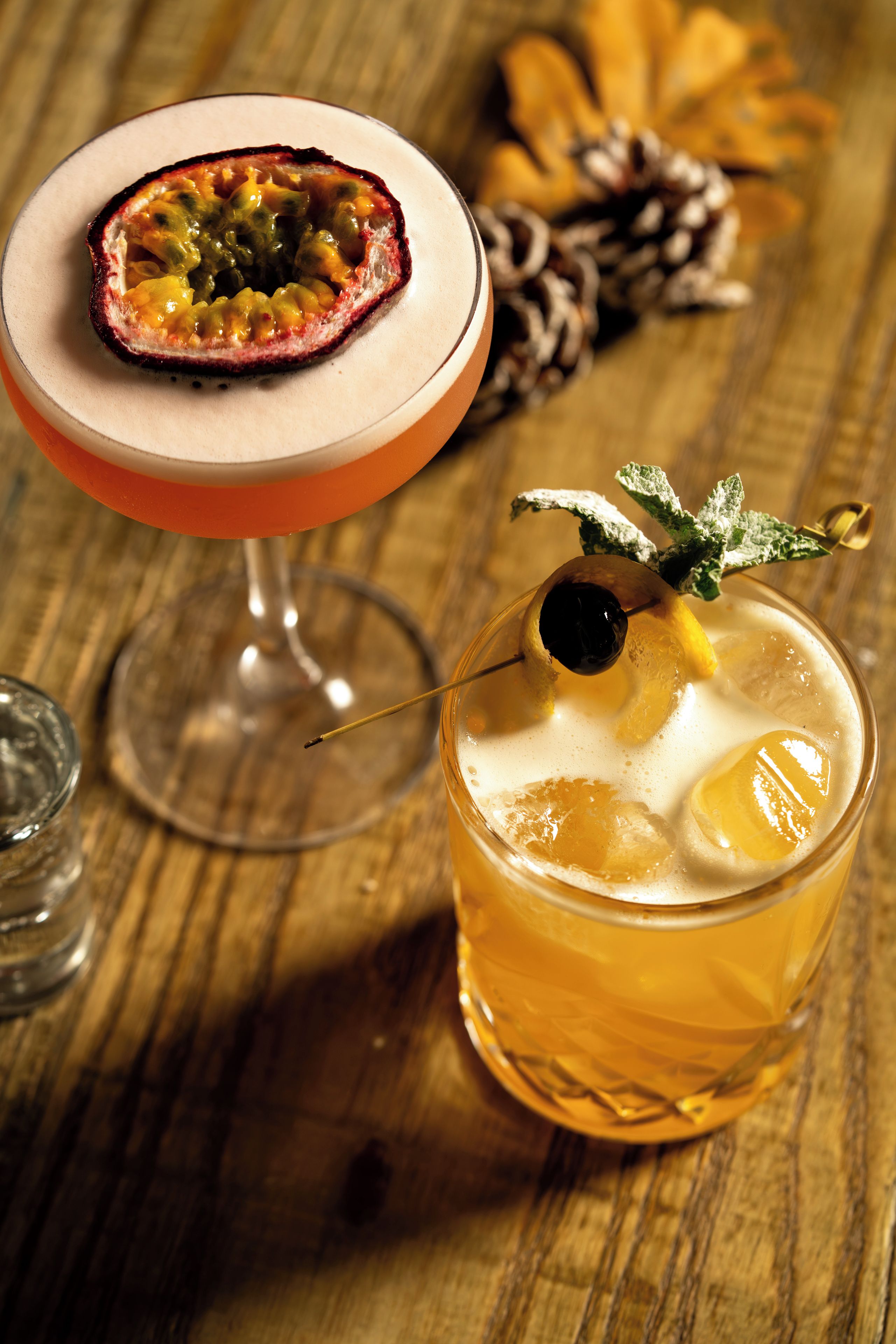 Due cocktail su un tavolo di legno, uno guarnito con frutti della passione e l'altro con una foglia di menta e una ciliegia.