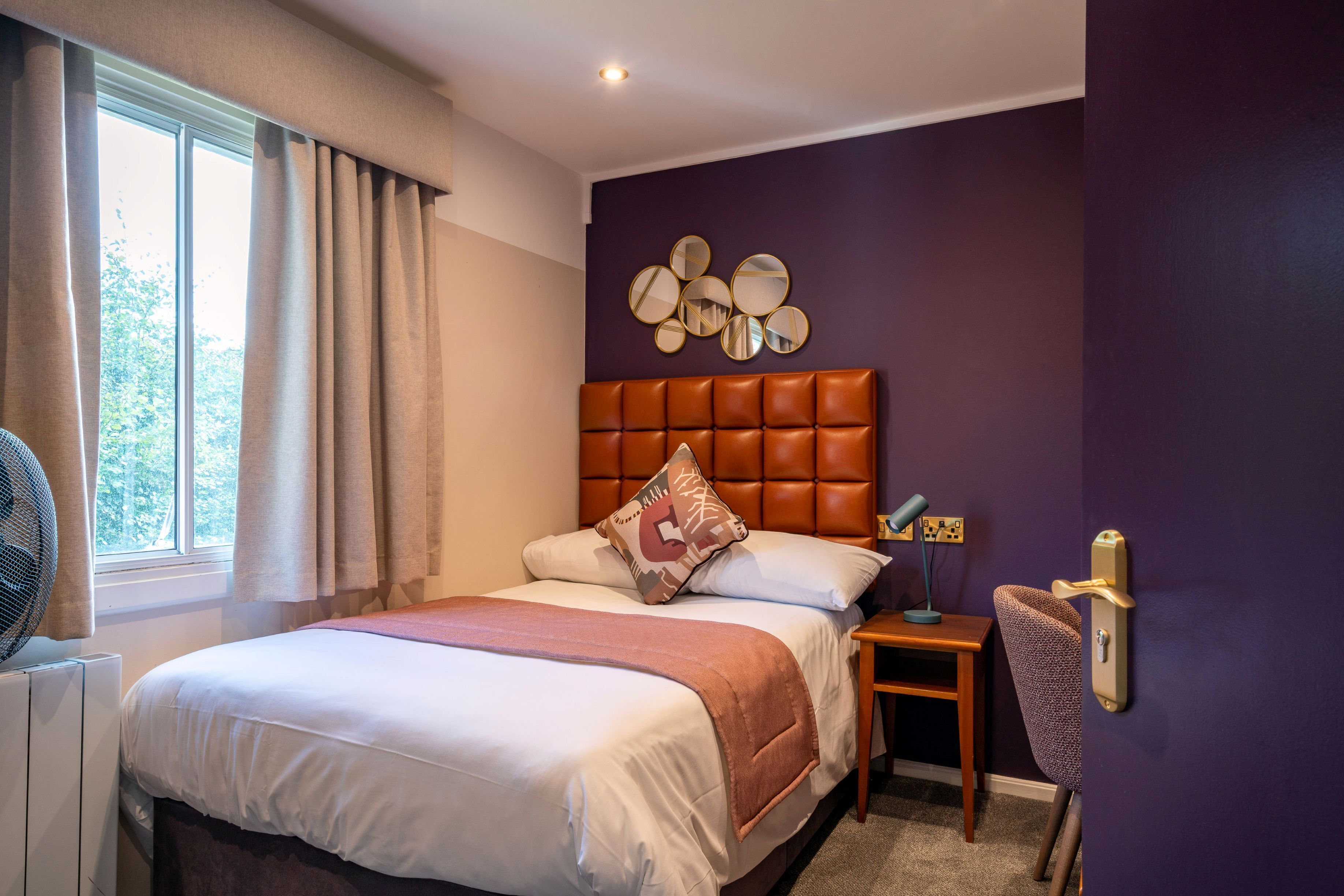 Una habitación de hotel con una cama grande, una mesita de noche con lámpara, un espejo en la pared y una ventana con cortinas.