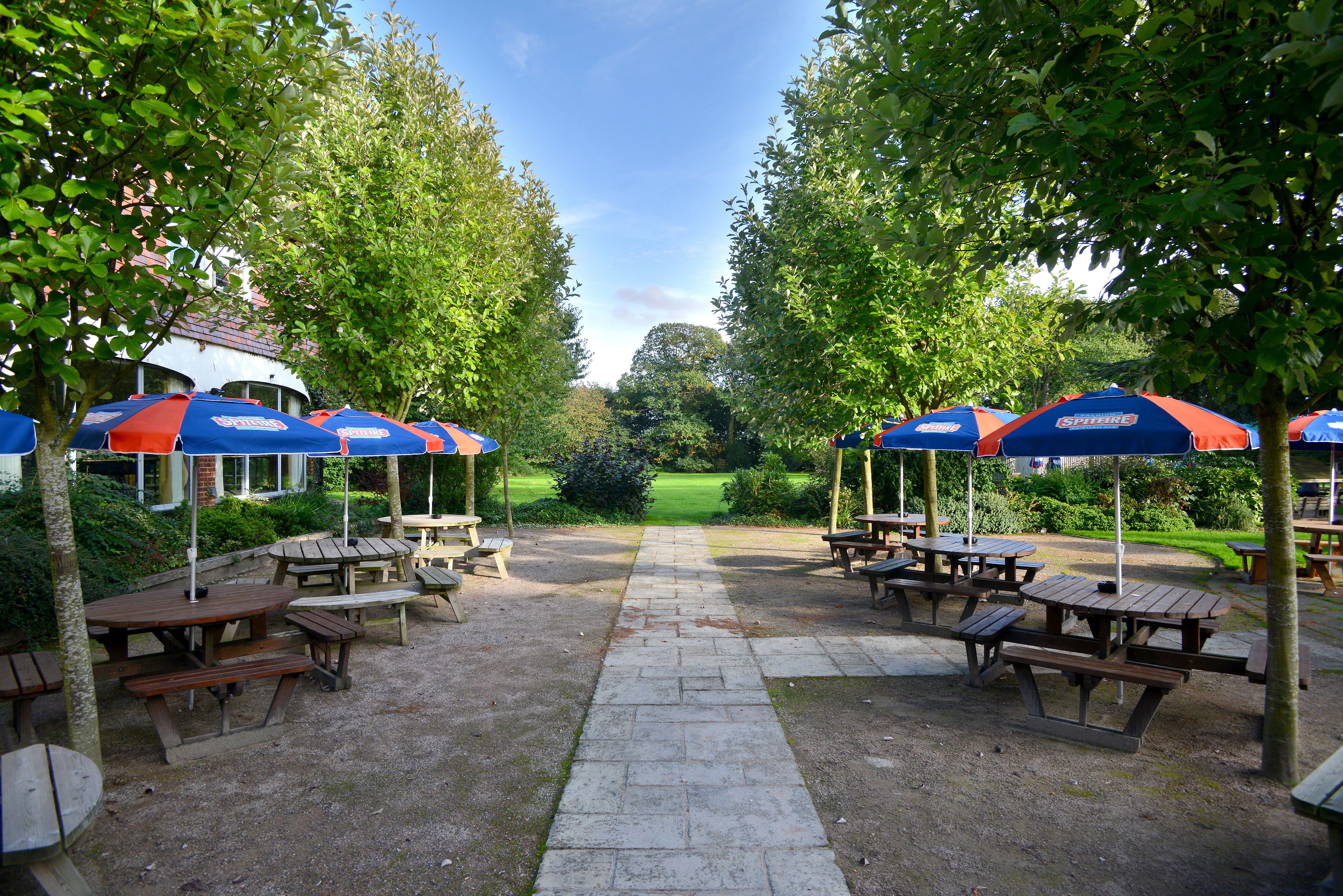 An outdoor patio area with picnic tables and umbrellas, surrounded by trees and a view of a golf course.