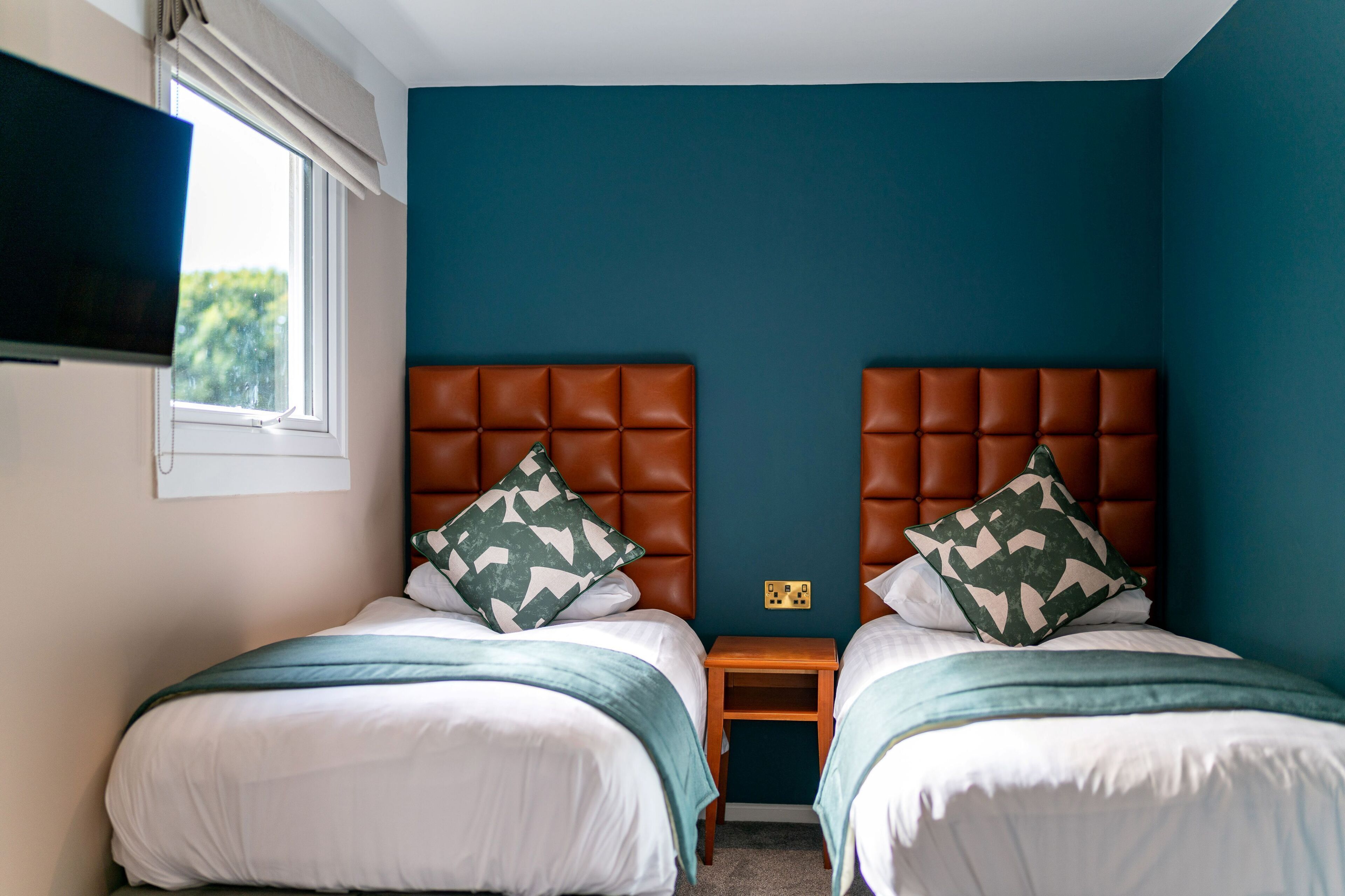A bedroom with two beds, each with a brown headboard, white bedding, and green throw pillows. There is a small wooden nightstand between the beds, and a TV mounted on the wall to the left.