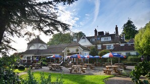 An outdoor seating area with tables and umbrellas, surrounded by greenery and a building with a traditional architectural style. - The Conningbrook Hotel (Ashford)