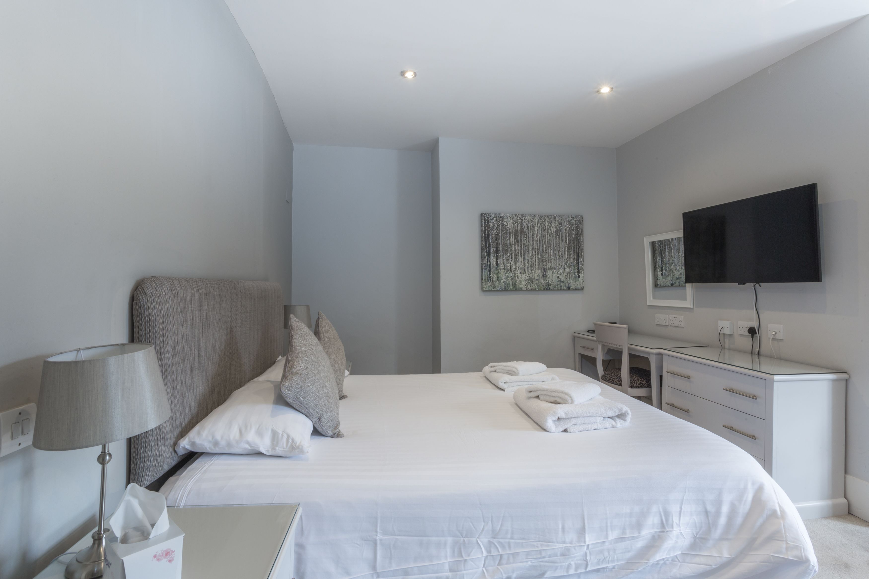A modern hotel room with a large bed, a bedside table with a lamp, a dresser, a flat-screen TV, and a painting on the wall.