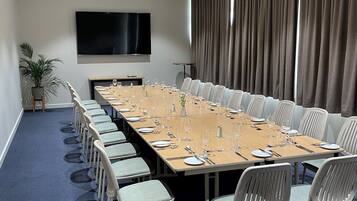 A conference room with a long table set for a meeting, surrounded by chairs, a flat-screen TV, and a large window with curtains.