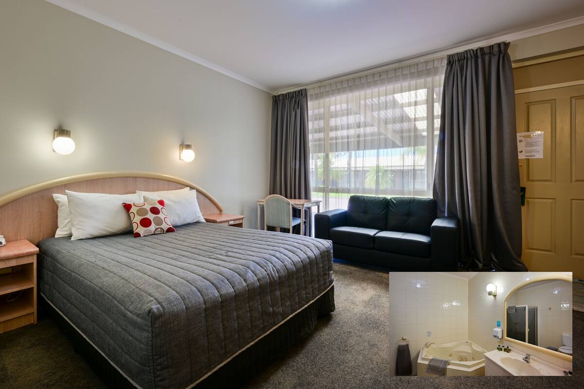 A hotel room with a large bed, bedside tables, a desk, a chair, a sofa, a window with curtains, and a bathroom with a bathtub and sink.