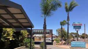 A hotel reception area with a sign for Westland Hotel Motel, featuring a reception desk, palm trees, and a parking lot. - Westland Hotel Motel (Whyalla Norrie)
