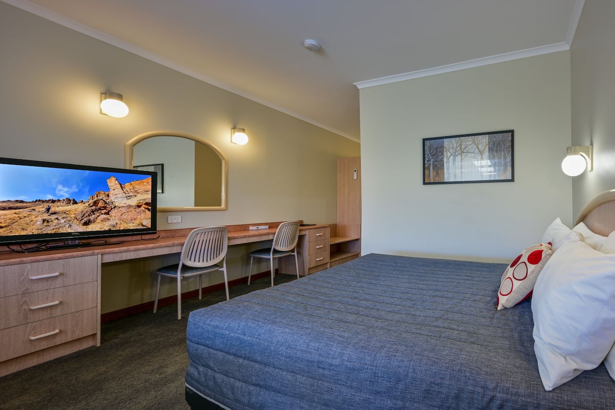 A hotel room with a bed, a desk with a chair, a television, and a wall-mounted light fixture.
