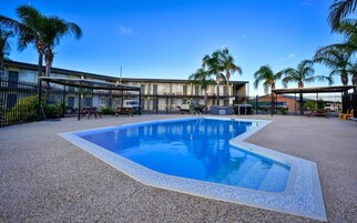 Piscina com borda azulejada, cercada por uma grade, palmeiras e um edifício com telhado plano.