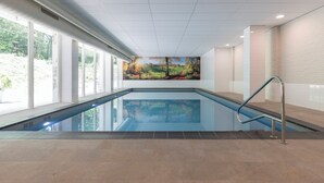 An indoor swimming pool with clear glass walls, a view of trees outside, and a mural on the wall. - Fletcher Hotel - Restaurant Erica (Berg en Dal)