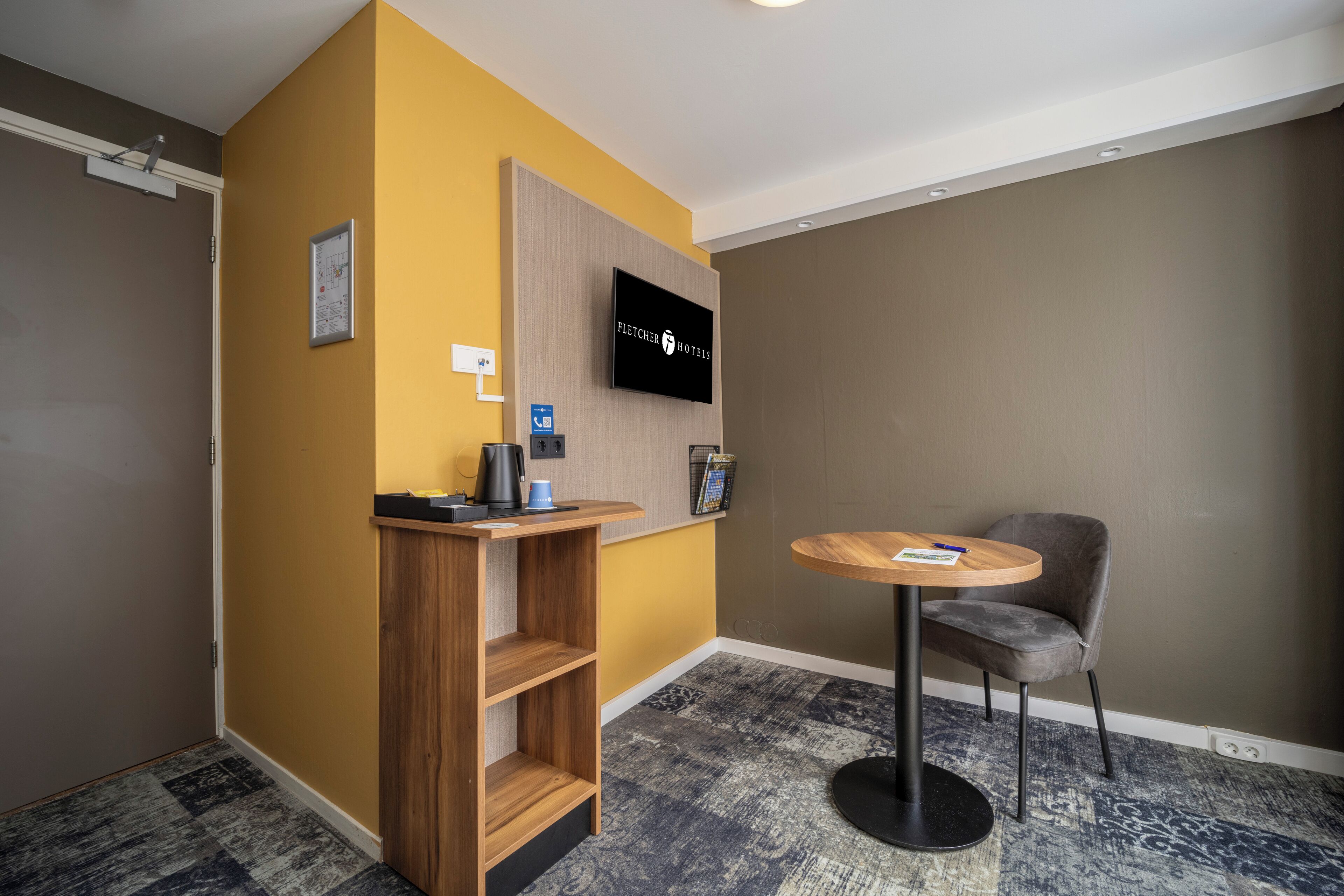 Une chambre d’hôtel avec un bureau en bois, une table ronde avec une chaise, un téléviseur fixé au mur et une étagère murale.