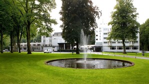 A hotel with a fountain in the center of a well-maintained lawn, surrounded by trees and a building with multiple windows and balconies. - Fletcher Hotel - Restaurant Erica (Berg en Dal)