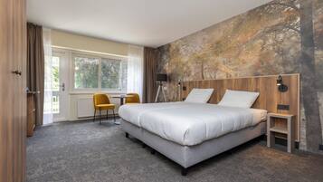 Una habitación de hotel moderna con una cama grande, un escritorio y una silla. Se observa una ventana con cortinas, una pared con un mural de un árbol y una mesita de noche con una lámpara.