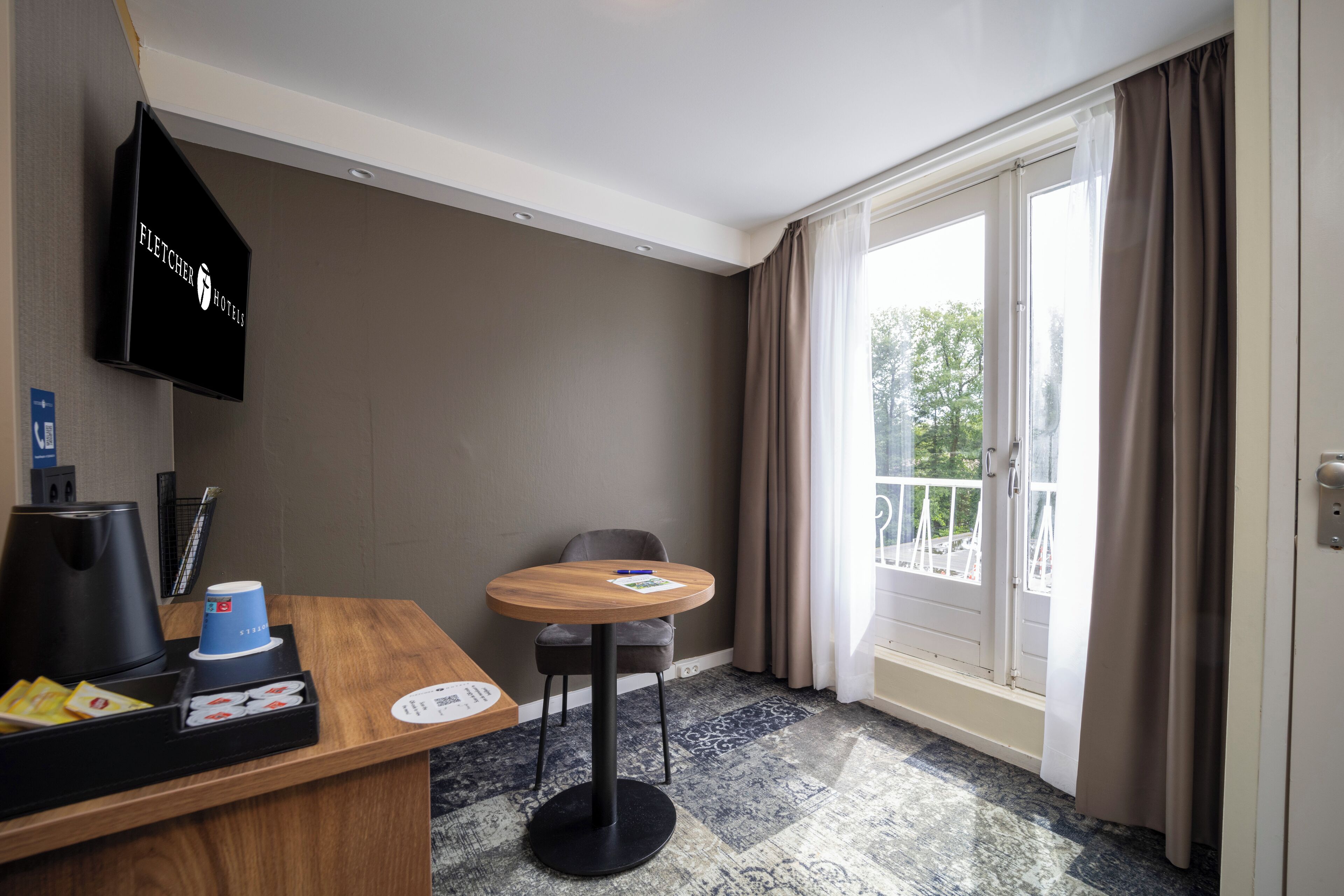 Une chambre d’hôtel avec un bureau en bois, une table ronde, une chaise, un téléviseur fixé au mur et une grande fenêtre avec des rideaux.