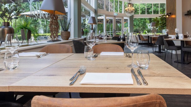 A restaurant with a wooden table set for two, large windows, and a chandelier. - Fletcher Hotel - Restaurant Erica (Berg en Dal)