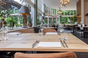 Un restaurant avec une table en bois dressée pour deux personnes, de grandes fenêtres et un lustre.