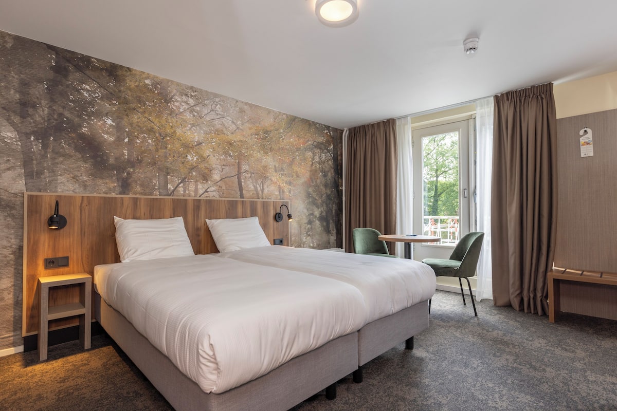 Hotellihuone, jossa on suuri sänky, pieni pöytä, kaksi tuolia, seinämaalaus ja ikkuna, jossa on verhot.