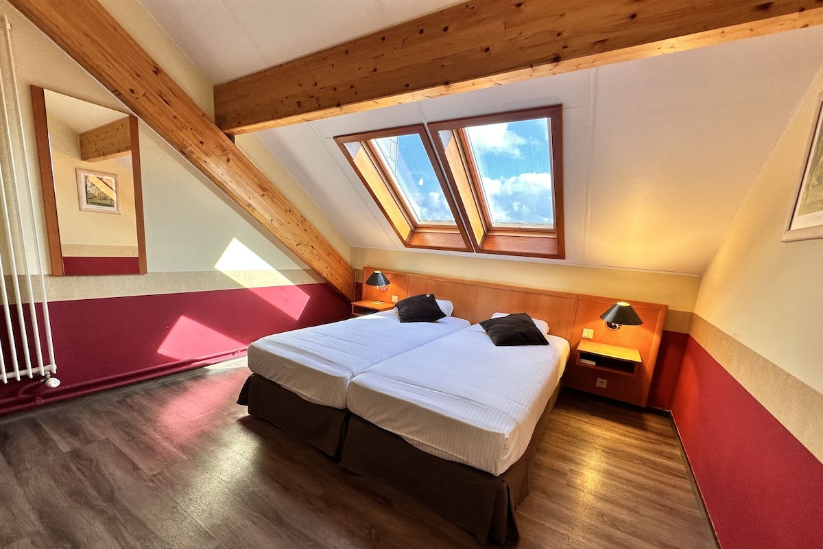 A bedroom with a sloped ceiling, wooden beams, a large bed with two pillows, a nightstand with a lamp, and a window providing natural light.