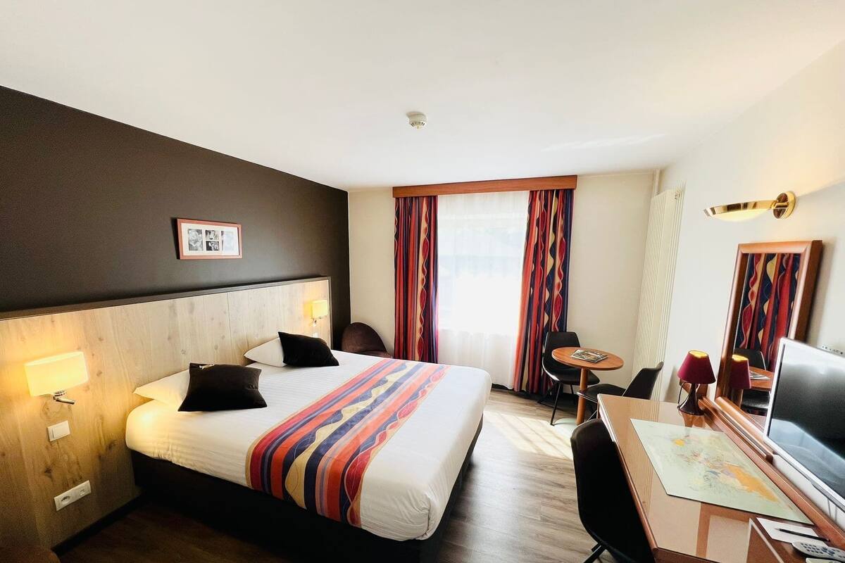 A hotel room with a bed, bedside lamps, a desk with a chair, a television, and a window with curtains.