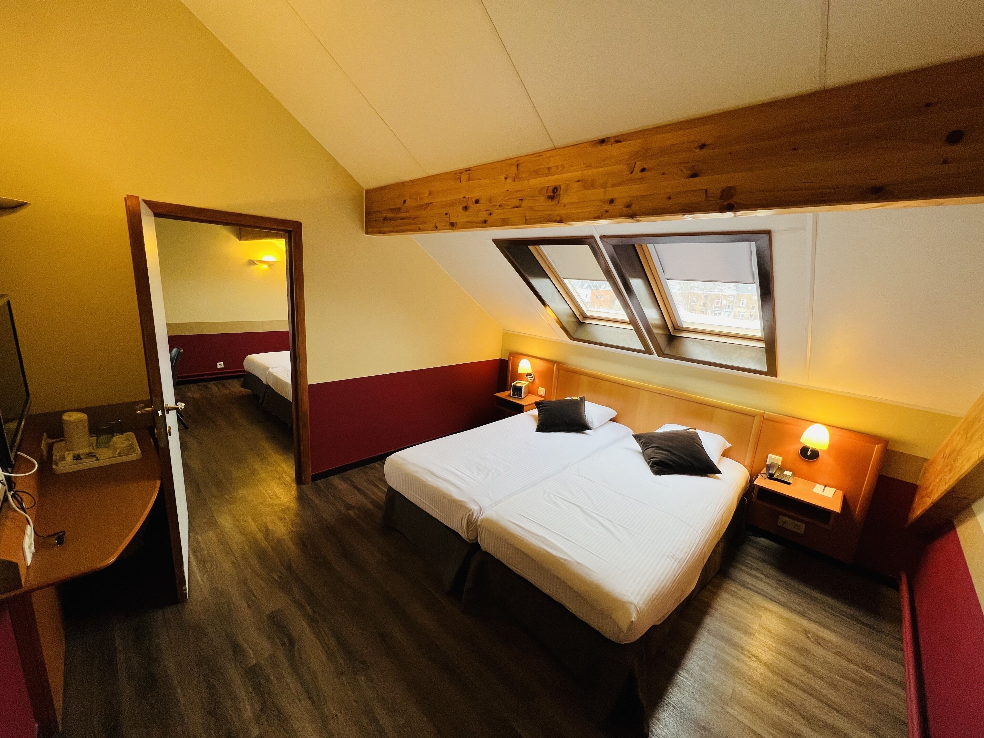 A hotel room with two beds, wooden ceiling beams, a skylight, and a small desk with a lamp.