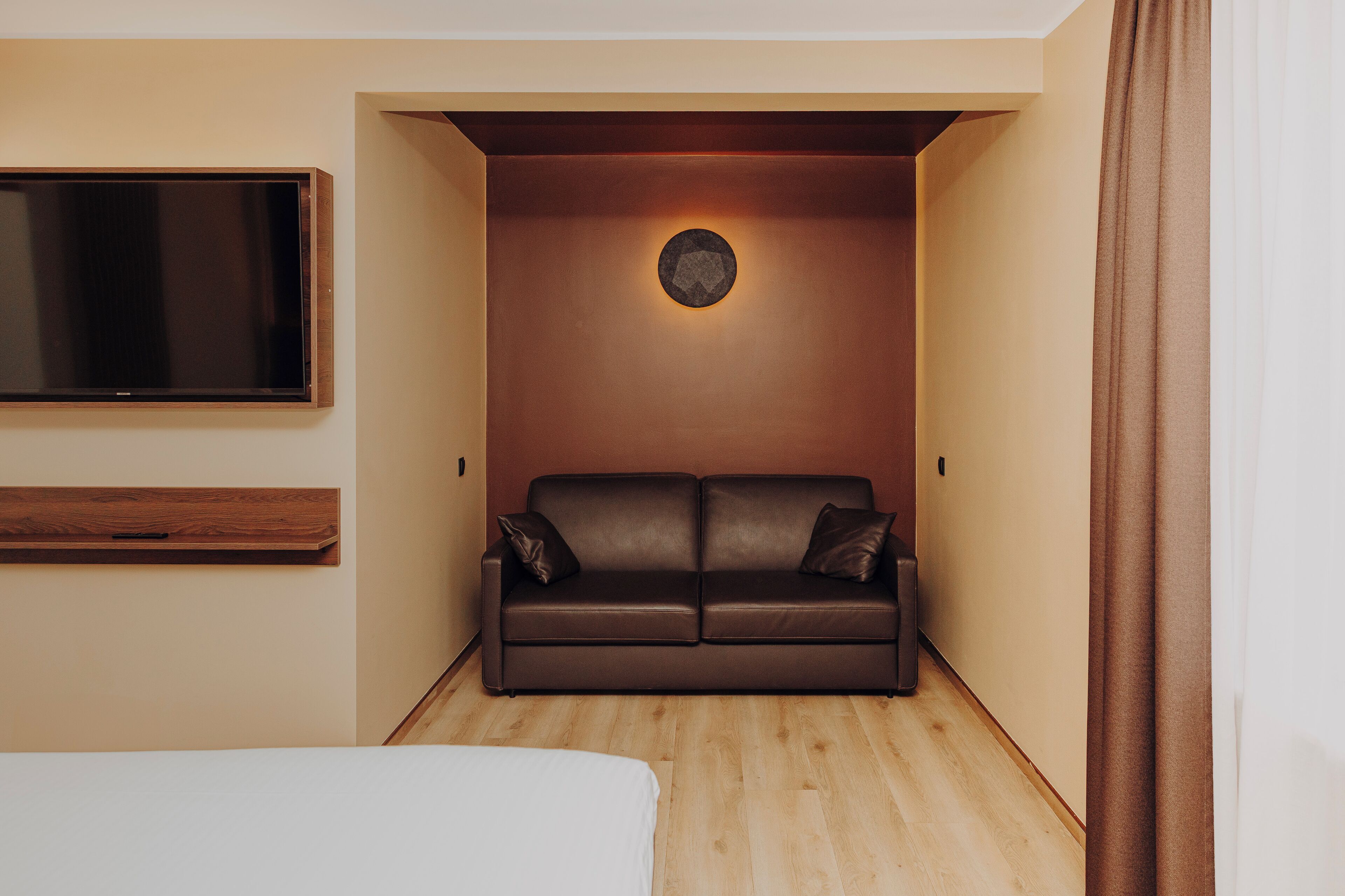 A modern hotel room with a brown leather sofa, a flat-screen TV mounted on the wall, and a wooden floor.