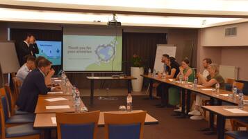 Sala de conferência com tela de apresentação, projetor e participantes sentados em uma mesa comprida com garrafas de água.