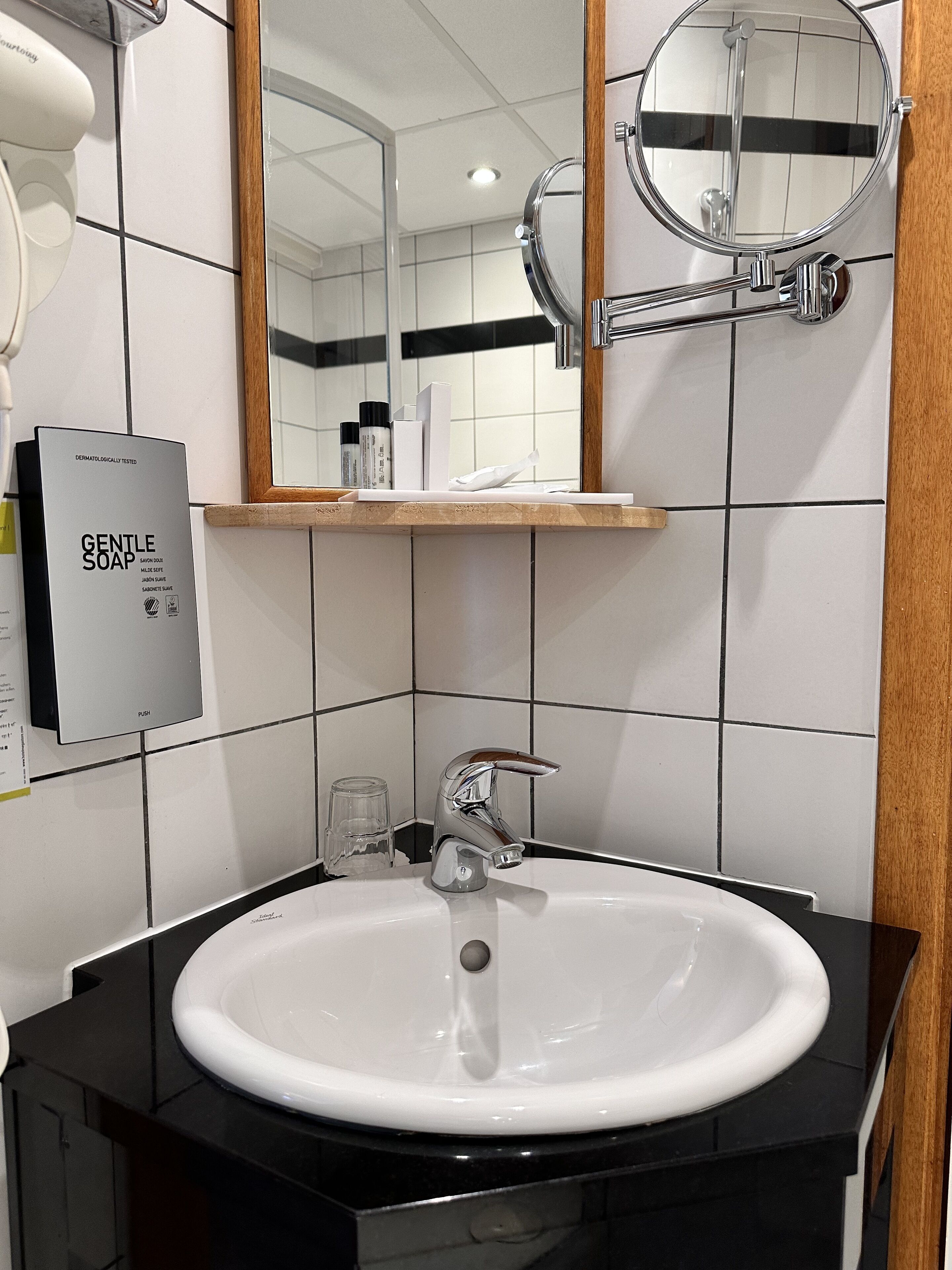 Un baño con lavamanos blanco, grifo cromado y un espejo encima. Hay un estante con artículos de aseo y un cartel que dice 'Jabón Suave'.