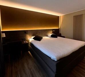 A hotel room with a large bed, a nightstand, a lamp, and a wall-mounted light fixture. - Hotel Melba (Bastogne)