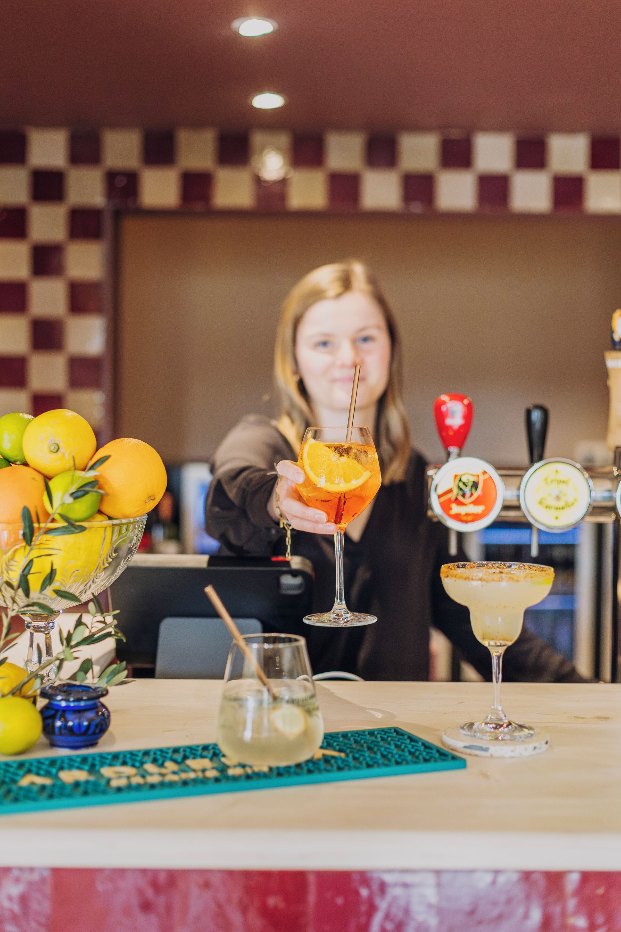 A bar area with a checkered wall, a bar counter, and a variety of drinks including a cocktail with an orange slice and a margarita.