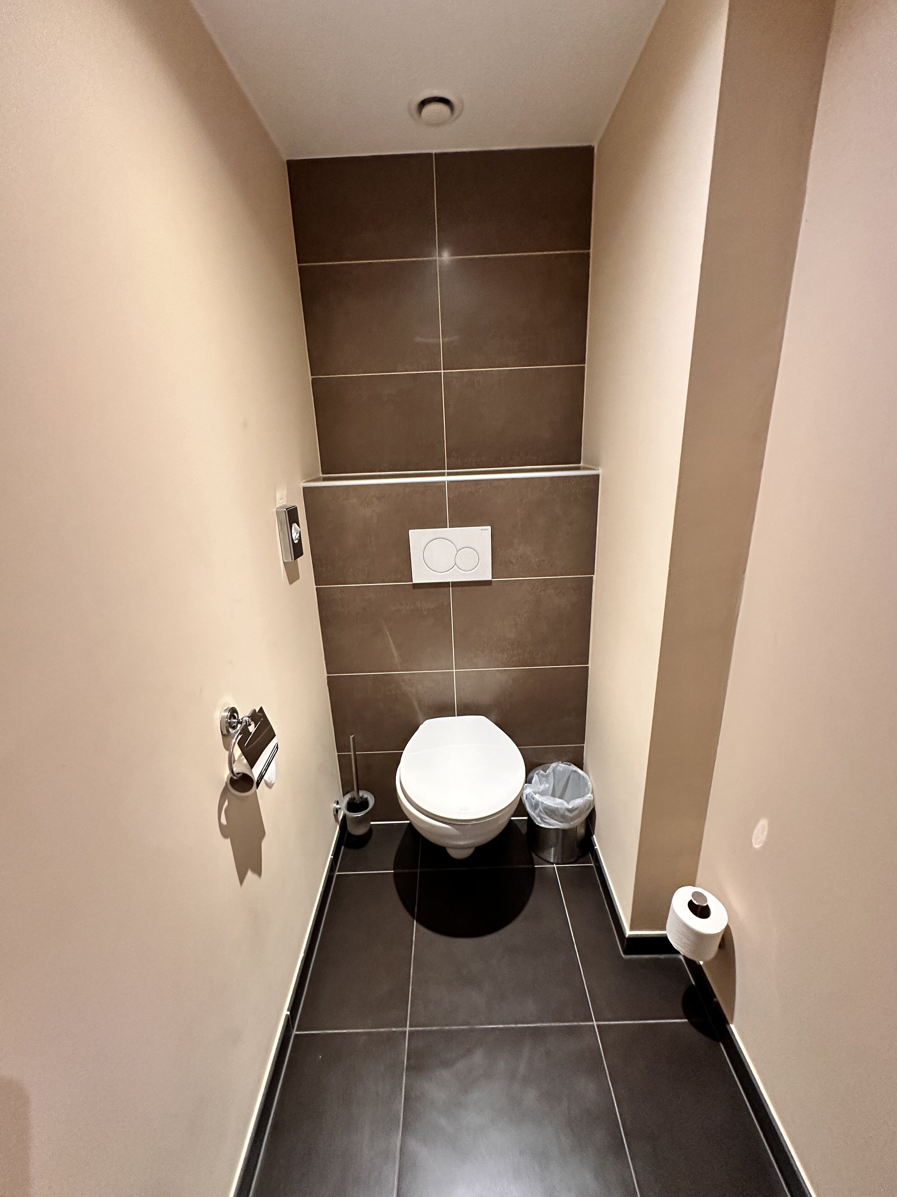 A small, modern bathroom with a toilet, a trash bin, and a toilet paper holder.